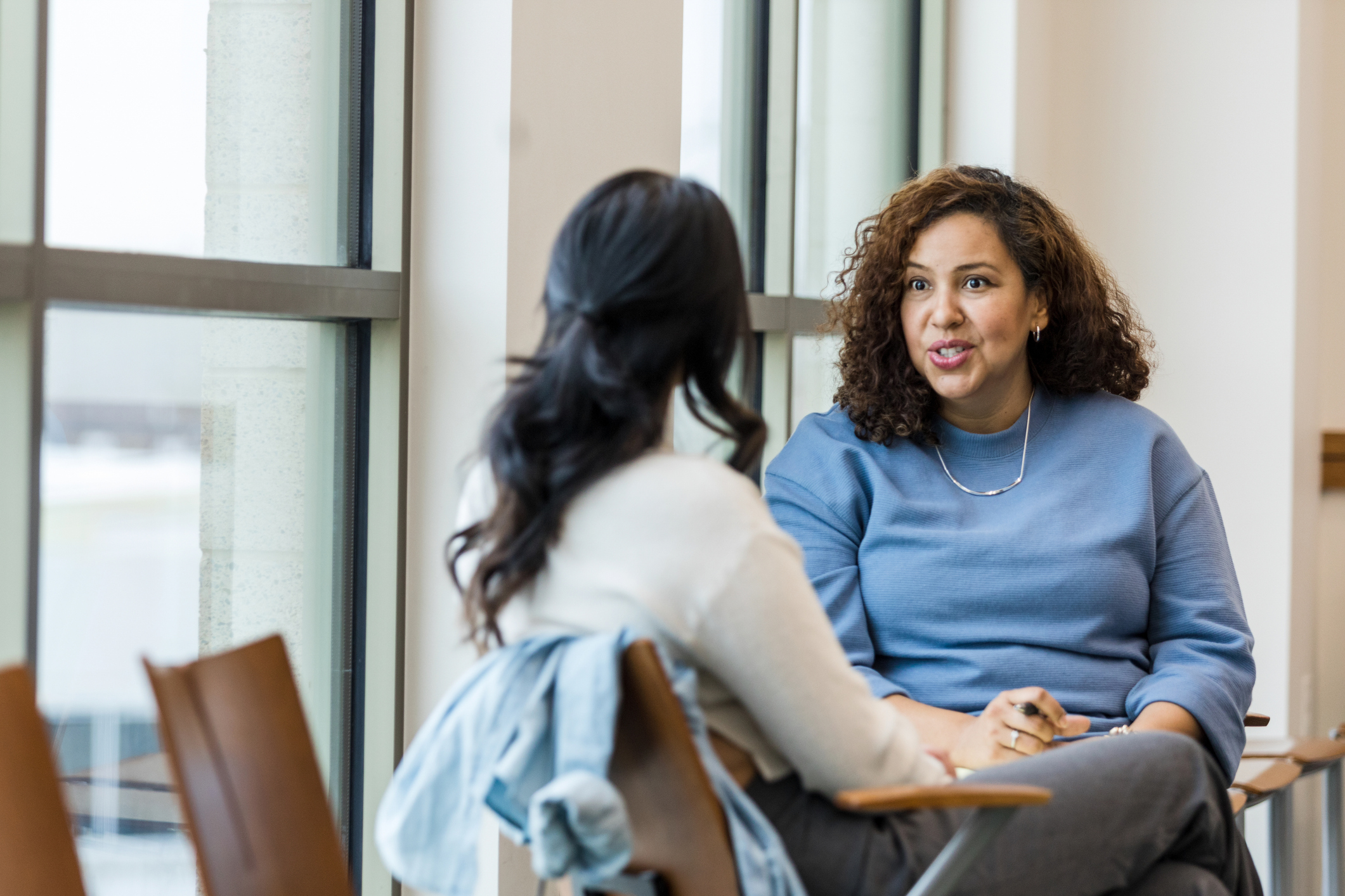 Two women sit near a window and talk.
