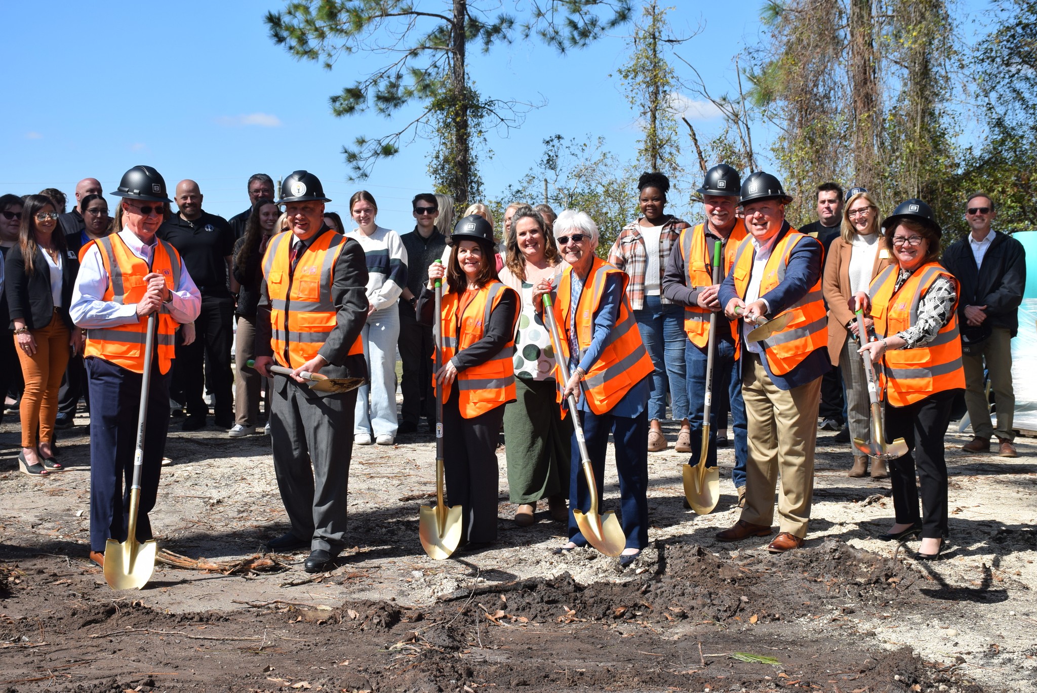 Seven people wearing orange vests and black hard hats, holding shovels in a groundbreaking ceremony. Croud of spectators behind them.