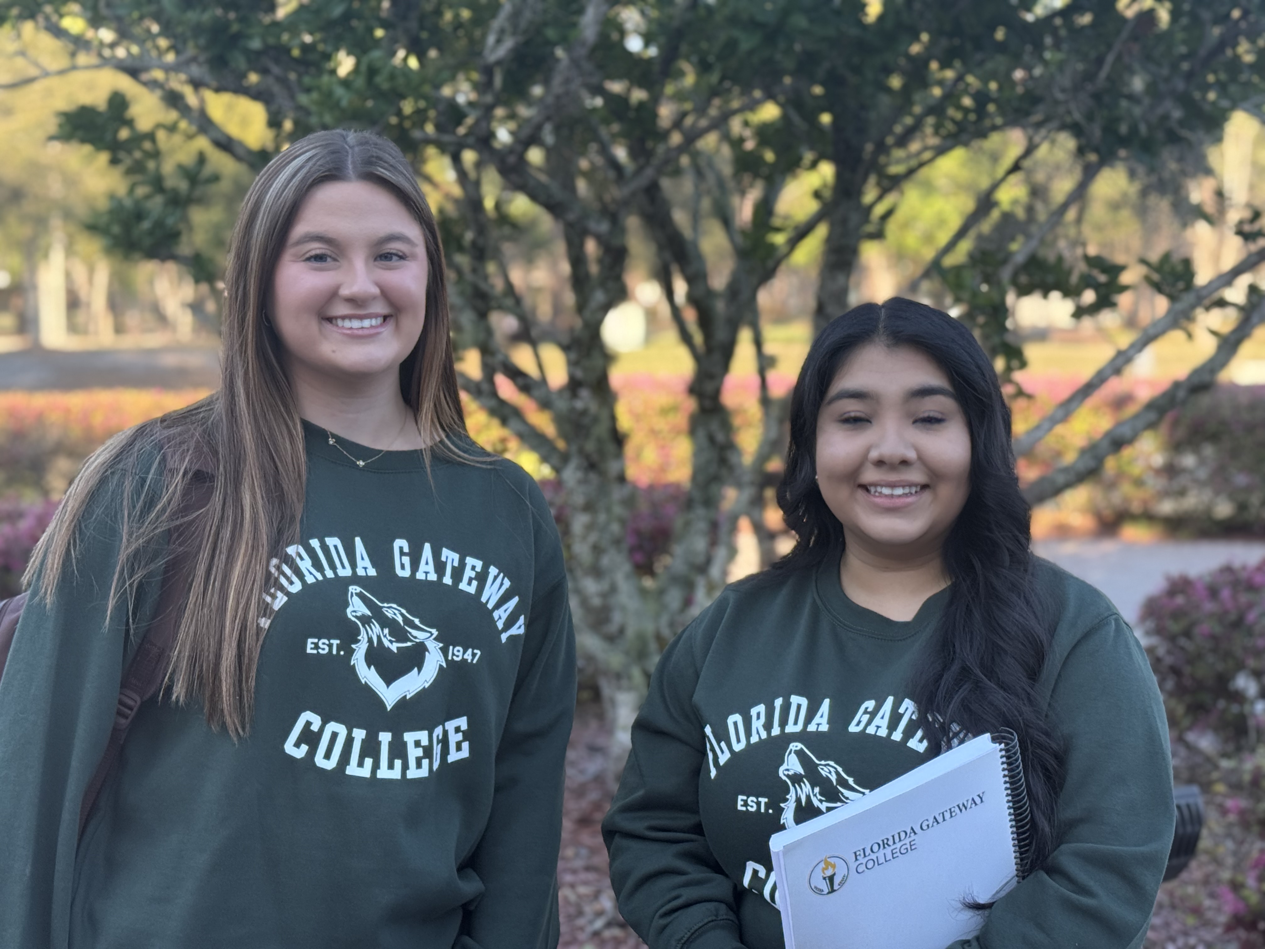 Two female students in FGC Sweatshirts smiling in front of tree