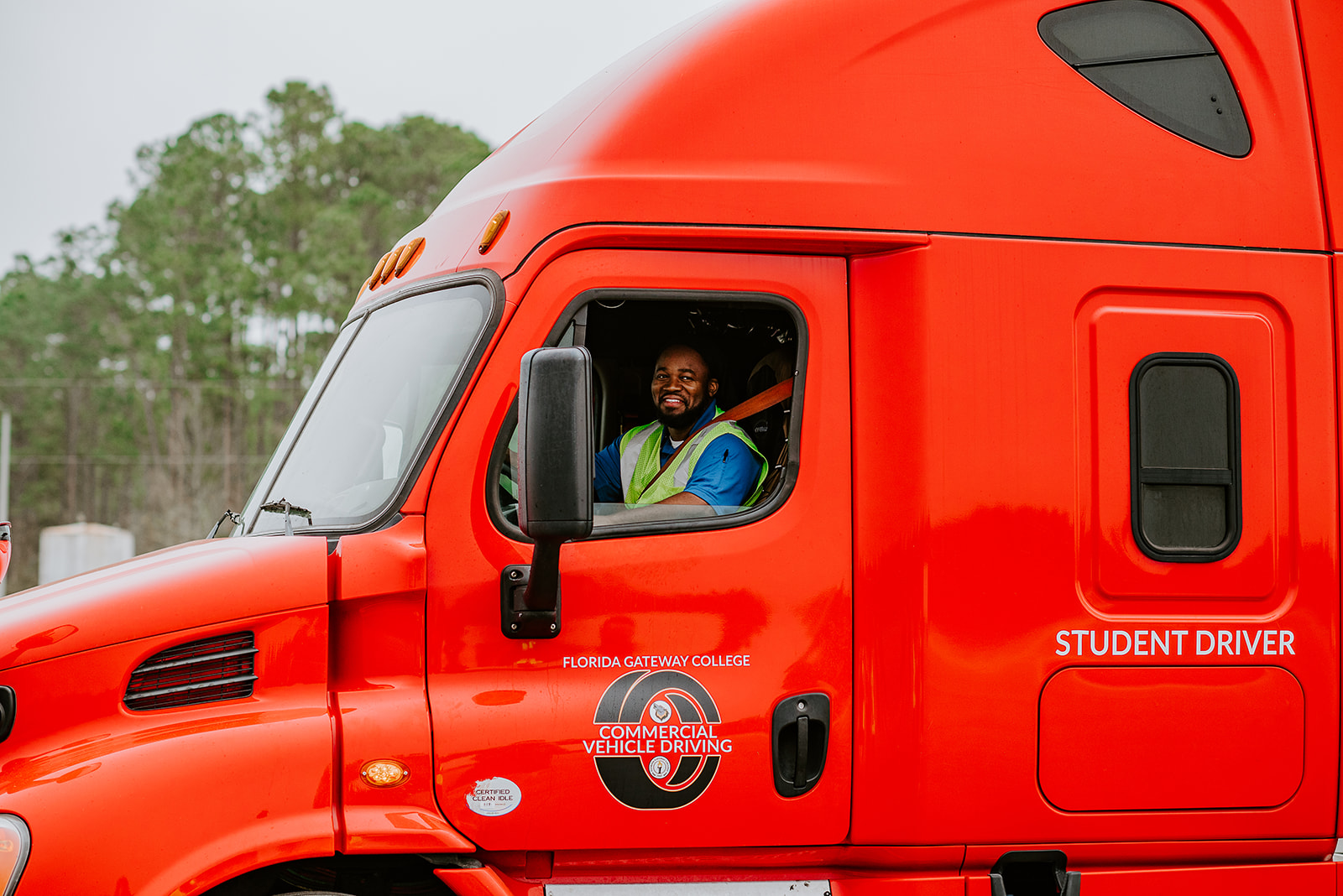 Student in yellow high-visibility vest sitting in cab of red tractor trailer.