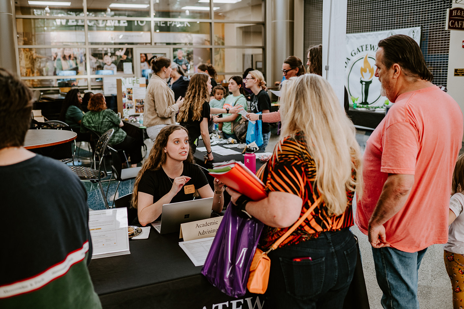 A woman at an academic advising table speaks with visitors at a busy college fair.