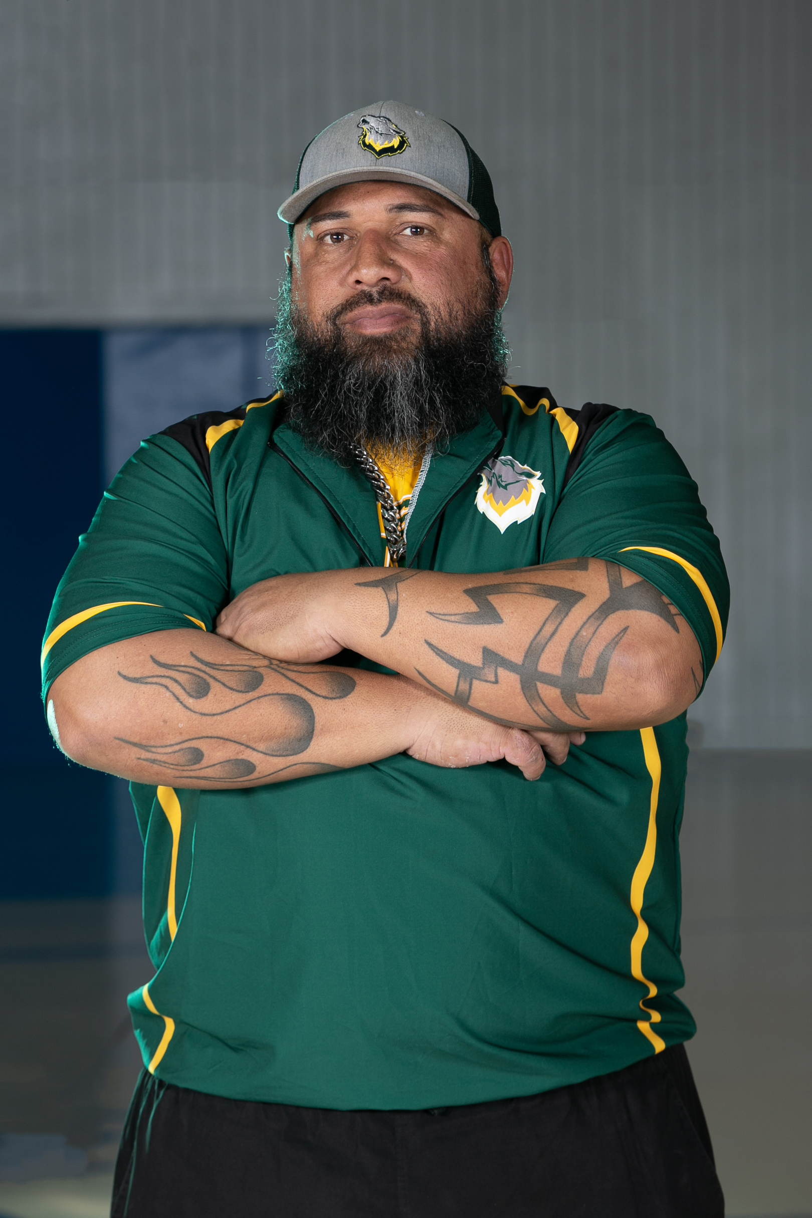 Portrait photo of FGC Softball Coach Jesus Rivera in a hat and green shirt with arms folded.