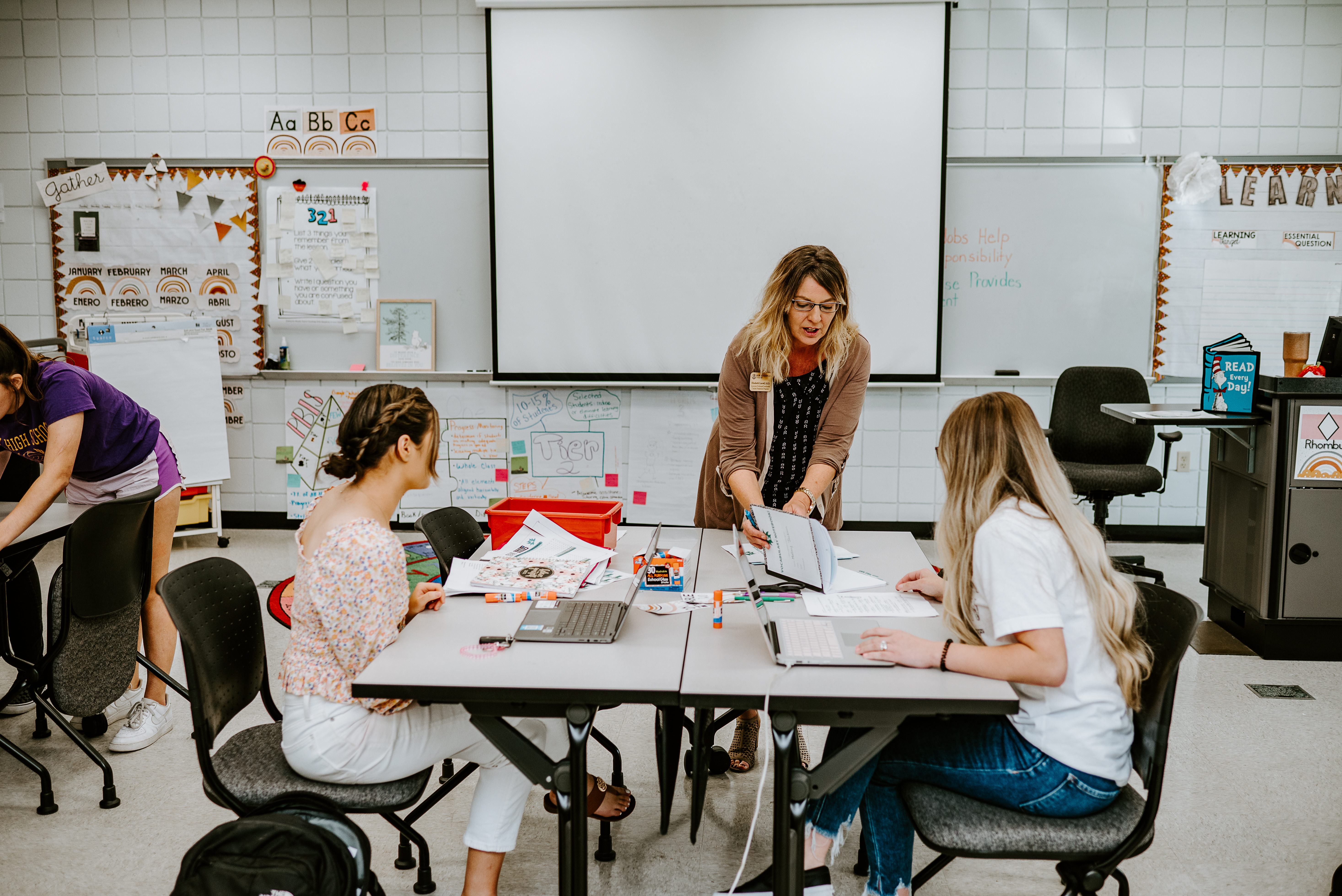 Instructor and students work together in a classroom, reviewing materials and collaborating at tables with laptops and teaching resources.