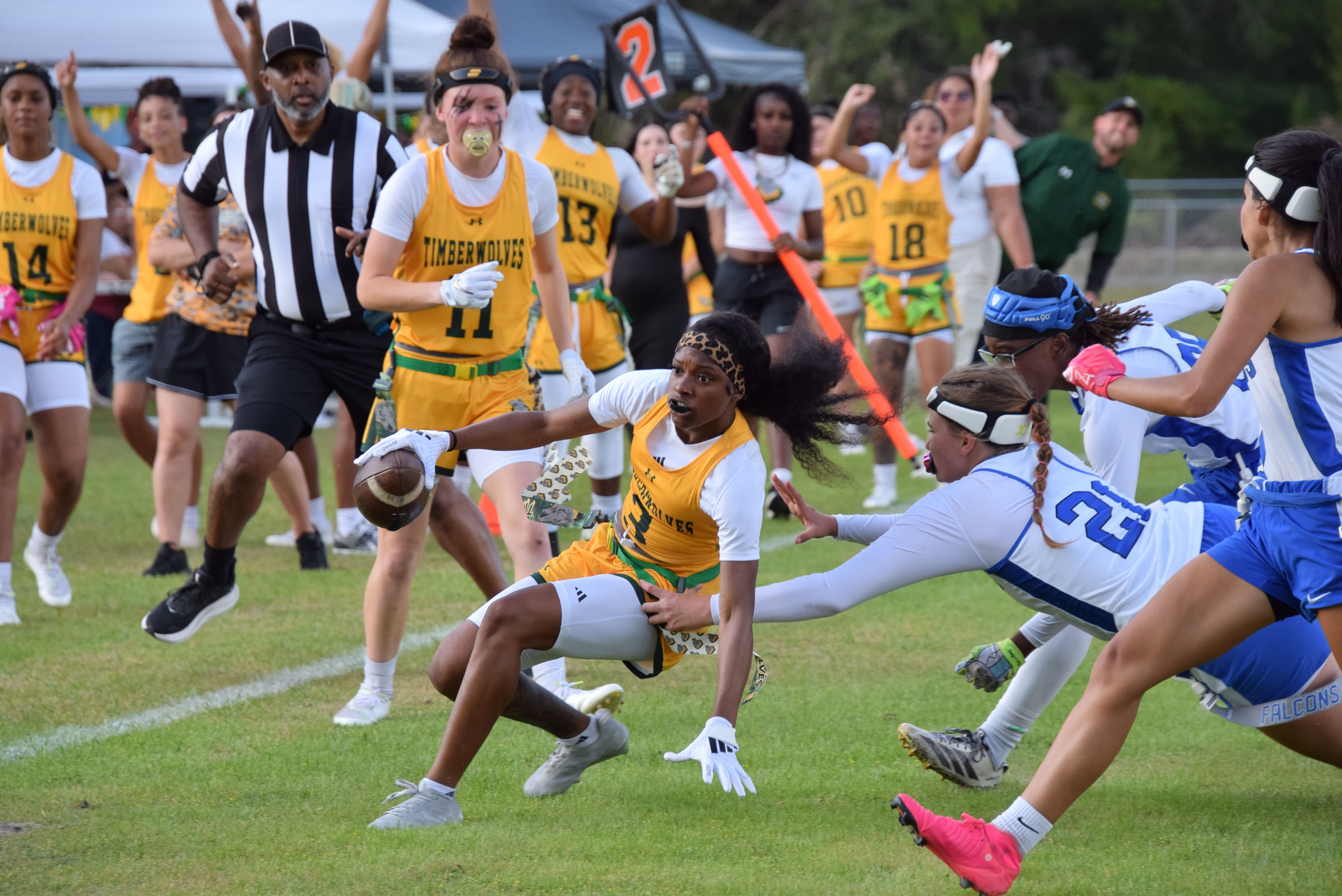 Women’s flag football game in action as a player runs with the ball while defenders reach to pull her flag, with teammates and a referee visible in the background.