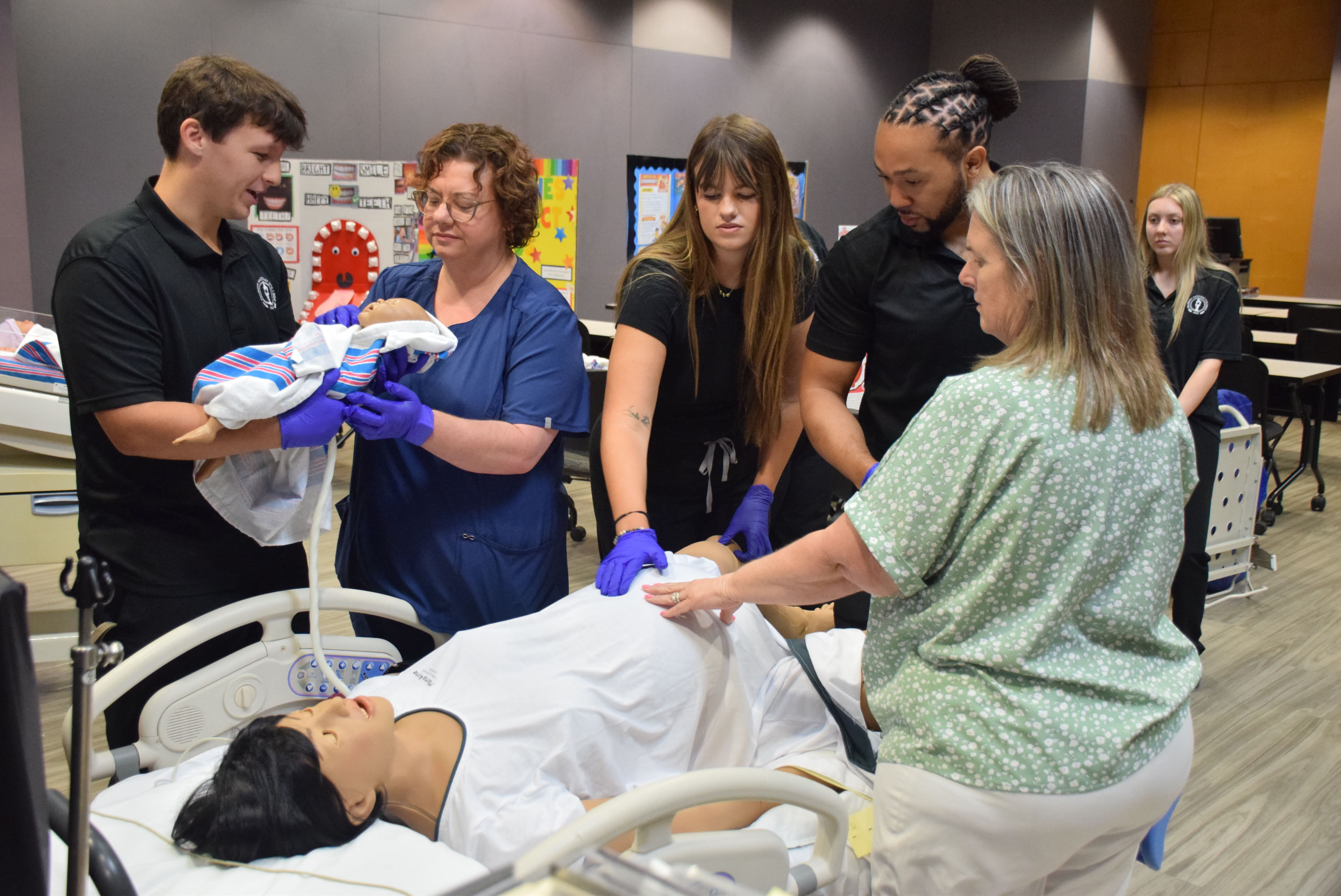 Nursing students and instructors participate in a hands-on medical training exercise using patient and infant simulation mannequins in a classroom lab.